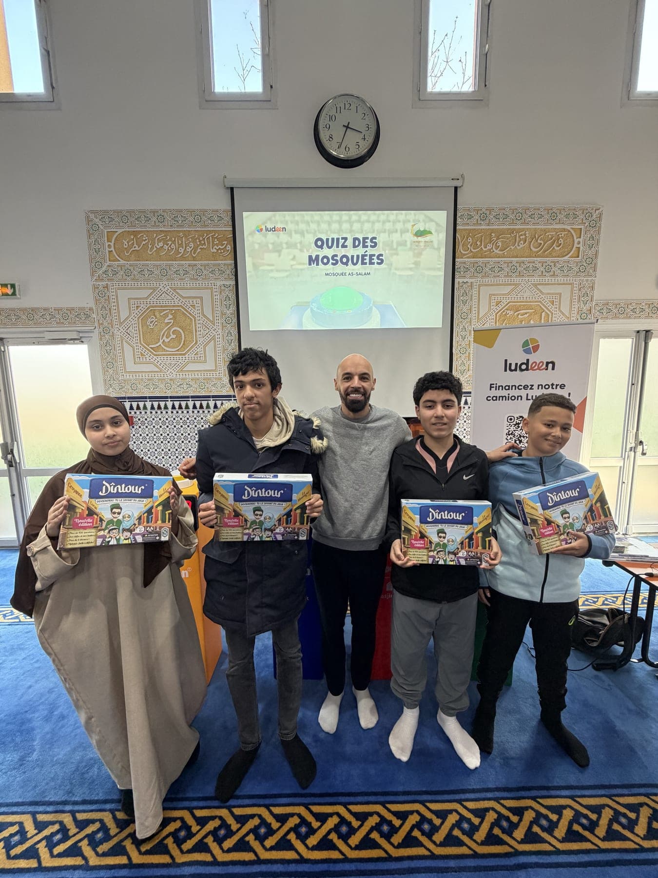 Quatre jeunes lauréats posant avec le jeu de société qu’ils ont remporté, devant l’écran du Quiz des Mosquées